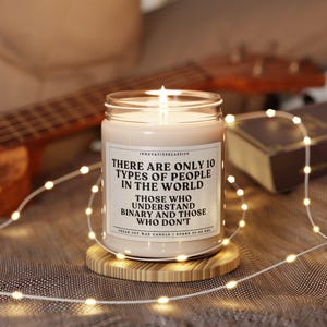 May include: A white soy wax candle with a black label that reads "There are only 10 types of people in the world. Those who understand binary and those who don't." The candle is sitting on a wooden coaster.
