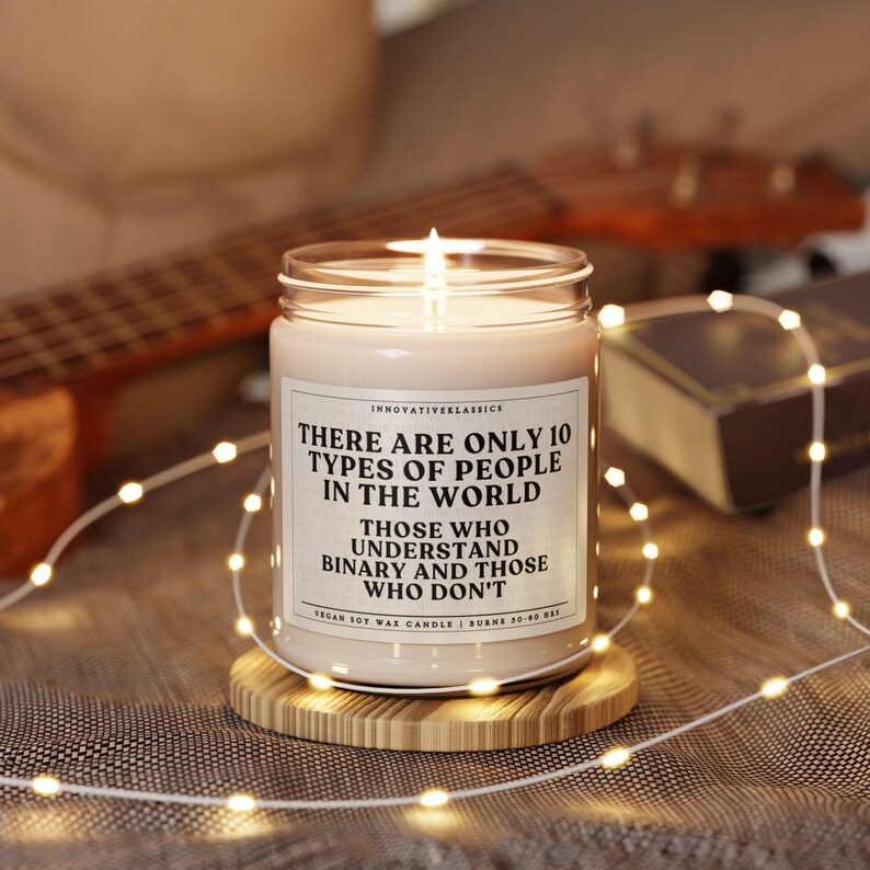 May include: A white soy wax candle with a black label that reads "There are only 10 types of people in the world. Those who understand binary and those who don't." The candle is sitting on a wooden coaster.