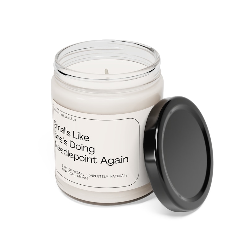 May include: A white candle in a clear glass jar with a black lid. The label reads "Smells Like She's Doing Needlepoint Again". The candle is 3 oz, vegan, completely natural, and made with non-toxic aromas.