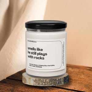 May include: White candle jar with a black lid. The label reads "smells like he still plays with rocks" and "8 oz of vegan, completely natural, non-toxic aromas".