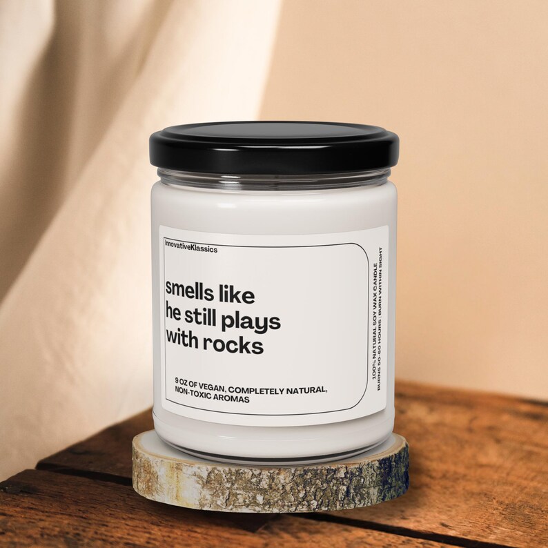 May include: White candle jar with a black lid. The label reads "smells like he still plays with rocks" and "8 oz of vegan, completely natural, non-toxic aromas".