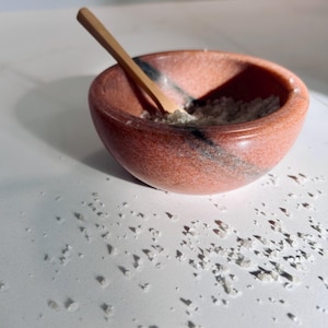 Könnte beinhalten: Eine kleine Schale aus rosa und schwarzem Marmor, gefüllt mit weißen Salzkristallen und einem Holzlöffel, der auf dem Rand liegt.