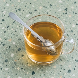 Puede incluir: Una taza de cristal transparente llena de té de color ámbar, con un infusor de té de acero inoxidable de mango largo sumergido en el líquido. El infusor tiene un cabezal ovalado perforado diseñado para infusionar té en hebras.
