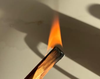 Puede incluir: Un palo santo encendido con una llama naranja. La madera es de color marrón claro con un extremo carbonizado oscuro. El fondo es de color neutro con sombras.