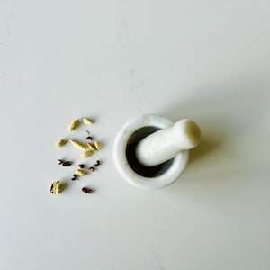 以下が含まれることがあります： 白い大理石のすり鉢とすりこぎセット。カルダモンの鞘と赤いペッパーコーンが添えられています。すり鉢は丸く、カーブしたハンドルが付いており、すりこぎは円筒形です。スパイスは白い表面に散らばっています。