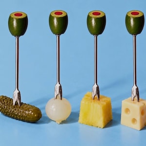 Puede incluir: Cuatro palillos de cóctel de metal plateado, cada uno rematado con una aceituna verde con un centro rojo. Los palillos están insertados en un pepinillo verde, una cebollita blanca, un cubo de piña amarillo y un cubo de queso amarillo.