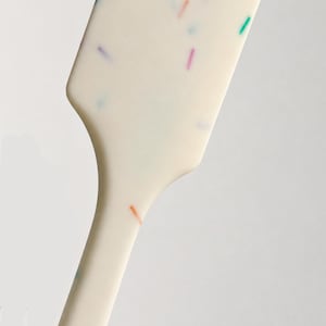 以下が含まれることがあります： ピンク、オレンジ、イエロー、ブルー、グリーンの小さな多色の斑点が散りばめられた、オフホワイトのキッチン用スパチュラ。平らで幅の広いヘッドが細いハンドルへとつながっており、滑らかな質感に仕上げられています。