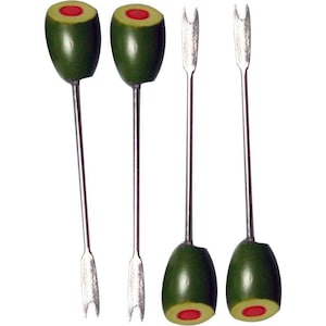 May include: A set of four metal cocktail picks, each featuring a green olive topper with a red pimento centre. The picks have a long, slender silver-toned shaft and a two-pronged fork at the bottom end for spearing garnishes in drinks.