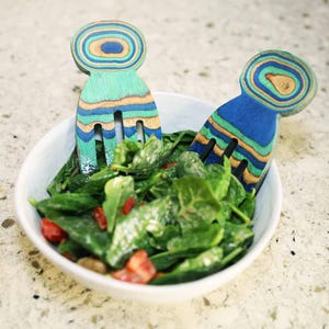 May include: A white bowl of fresh spinach salad with red tomatoes. Two decorative salad servers with a layered, colourful wood design are in the bowl. The salad servers are ready to serve the salad.