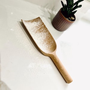 Puede incluir: Una cuchara de madera espolvoreada con harina blanca, que se encuentra sobre una encimera blanca. La cuchara tiene un mango curvo y un cuenco ancho y poco profundo.