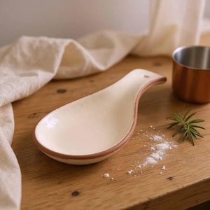 Puede incluir: Un reposacucharas de cerámica con un interior liso de color crema y un borde en contraste de color terracota. El reposacucharas presenta una forma ovalada alargada y un pequeño orificio en el extremo del mango para colgarlo, diseño sencillo.