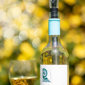 May include: A clear glass wine bottle with a light blue foil neck and a clear plastic pourer spout inserted into the top. The white label features a teal wave graphic and the text La Révérence, Sauvignon Blanc, 2019, Pays d'Oc. A wine glass sits nearby.
