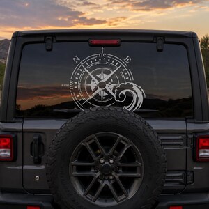 May include: A white vinyl decal applied to the rear window of a dark gray vehicle. The decal features a detailed nautical compass rose design with cardinal directions N, E, S, and W, accompanied by a stylized graphic of a crashing ocean wave.