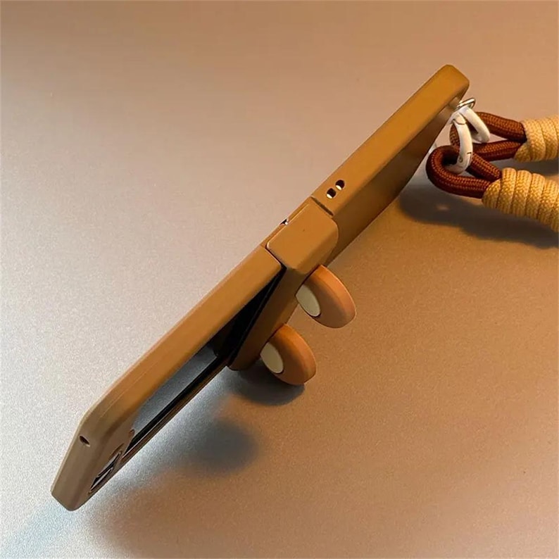 May include: A tan smartphone case with a side-mounted hinge mechanism featuring two small, round, tan and cream-colored decorative elements. A thick, braided cord strap in shades of brown and tan is attached to the top via a white metal clip.