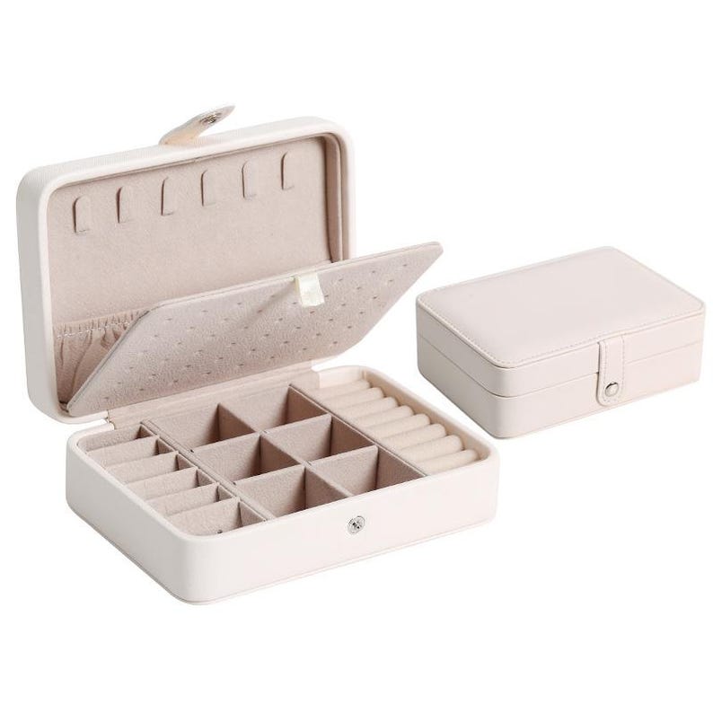 May include: A light pink rectangular jewelry box with a smooth exterior and a snap closure. The interior features a beige velvet lining with a hinged divider, multiple square compartments for small items, a ring roll section, and necklace hooks.