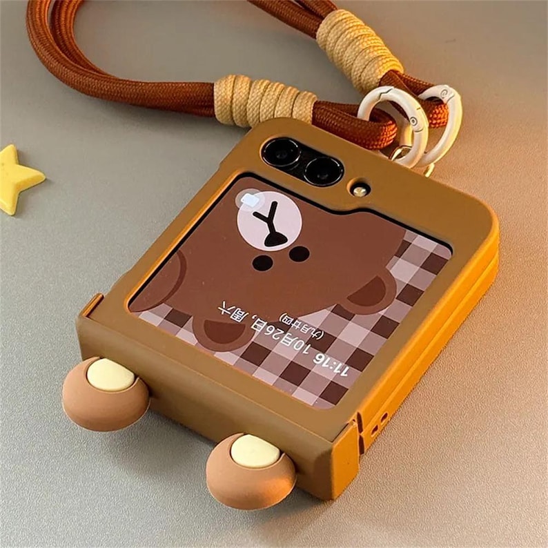 May include: A brown foldable phone case featuring two protruding bear ears at the bottom, a brown bear face graphic on the screen, and a brown woven lanyard attached with white clips. The screen displays the time 11:16 and the date October 26.