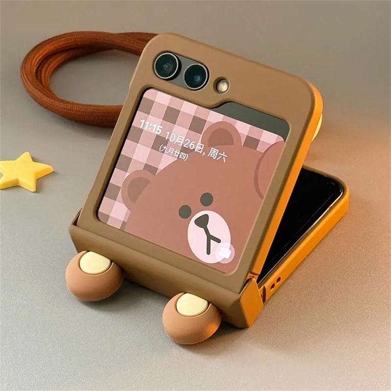 May include: A brown foldable phone case with two protruding circular accents at the bottom. The screen displays a brown bear graphic, a pink and brown checkered pattern, and the text 11:15 10月26日, 周六 (九月廿四). A brown strap is visible in the background.