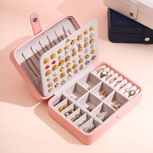May include: A light pink rectangular jewelry box with a snap closure. The interior features a hinged panel with multiple holes for earrings and a main compartment divided into small sections for rings and various accessories, lined in light gray.