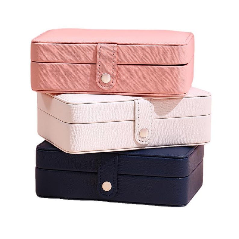May include: A stack of three rectangular jewelry boxes with a textured finish. The top box is light pink, the middle box is off-white, and the bottom box is navy blue. Each box features a central vertical strap with a silver-toned metal snap button.