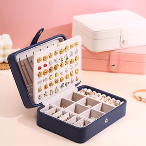 May include: A navy blue rectangular jewelry box with a hinged lid featuring a vertical panel for stud earrings and a base with multiple compartments for rings and small accessories, accompanied by two additional closed boxes in white and pink.
