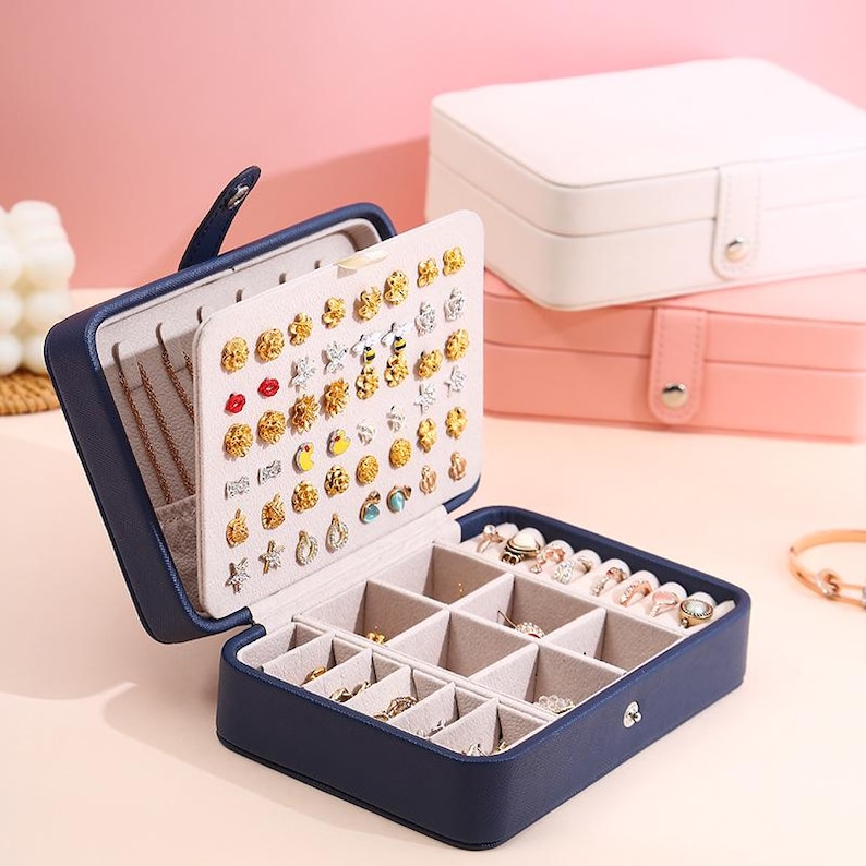 May include: A navy blue rectangular jewelry box with a hinged lid featuring a vertical panel for stud earrings and a base with multiple compartments for rings and small accessories, accompanied by two additional closed boxes in white and pink.