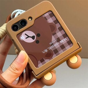 May include: A brown protective phone case for a foldable smartphone featuring a cutout for the camera lenses. The screen displays a brown bear graphic on a checkered background with the text 11:24 10月26日, 周六 (九月廿四). A brown wrist strap is attached.