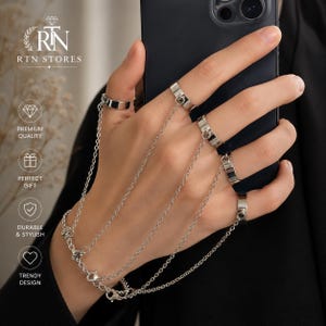 May include: A silver-toned hand harness featuring five plain metal rings connected by delicate link chains. The chains extend from each ring to a central wrist chain, creating a draped effect across the back of the hand. The jewelry is metallic.
