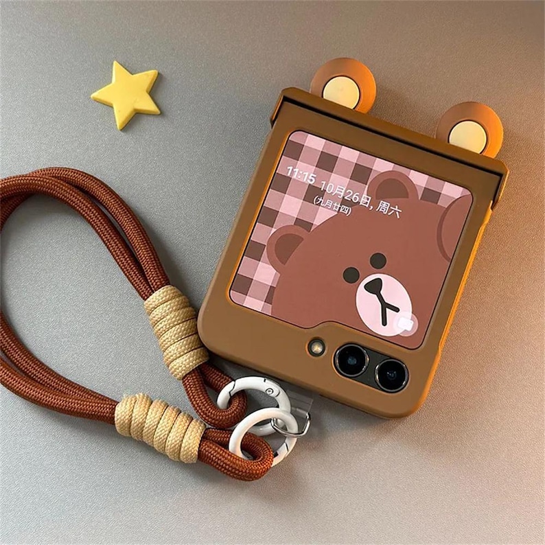 May include: A brown flip phone case with two rounded bear ears on top, featuring a screen display showing a brown bear graphic, a plaid background, and the text 11:15 10月26日, 周六 (九月廿四). A brown braided lanyard with beige accents is attached.