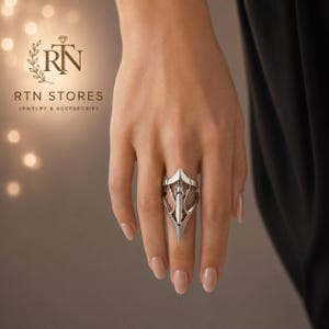 May include: A silver-toned metal ring featuring an elongated, pointed design that covers a significant portion of the finger. The ring has a structured, architectural shape with a central vertical element and open cutouts on the sides.
