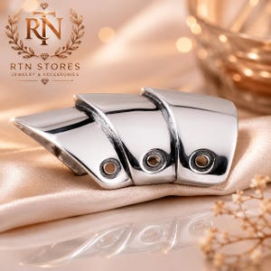 May include: A silver-toned, multi-layered armor ring featuring three articulated, polished metal segments. Each segment has a smooth, curved surface and a single, small circular cutout near the bottom edge, creating a sleek, segmented design.