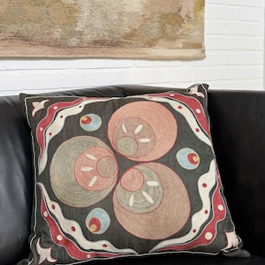 Könnte beinhalten: Dekoratives quadratisches Kissen mit einem gestickten Design. Das Kissen hat einen dunkelgrauen Hintergrund mit einem zentralen Muster aus sich überlappenden Kreisen in Pfirsich-, Grün- und Weißtönen. Der Rand ist rot und weiß mit einem sich wiederholenden Muster.