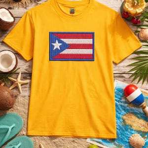 Peut inclure: Un t-shirt jaune avec un drapeau portoricain brodé. Le drapeau comprend un triangle bleu avec une étoile blanche et des rayures rouges et blanches. Le t-shirt est entouré d'articles sur le thème de la plage, comme un chapeau de paille, des lunettes de soleil et des noix de coco.