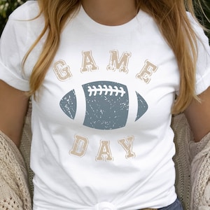 May include: White t-shirt with a distressed graphic of a football and the text "GAME DAY" in a vintage font.