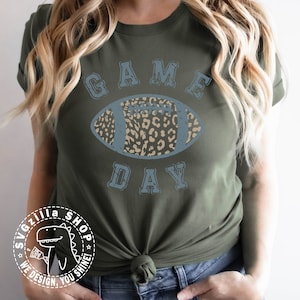 Pu&ograve; includere: Una maglietta verde con una grafica blu rovinata di un pallone da calcio con un design a stampa leopardata. Il testo "GAME DAY" &egrave; sopra il pallone.