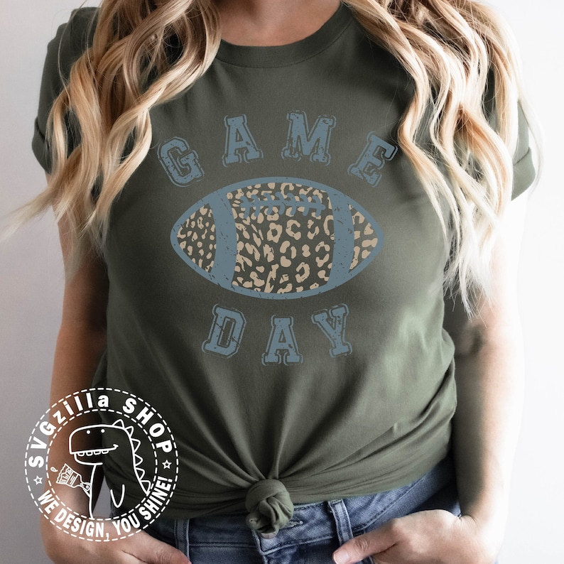 Pu&ograve; includere: Una maglietta verde con una grafica blu rovinata di un pallone da calcio con un design a stampa leopardata. Il testo "GAME DAY" &egrave; sopra il pallone.