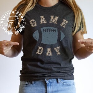 May include: A dark grey t-shirt with the text "GAME DAY" in a vintage style font. A distressed blue football graphic is centred on the shirt.
