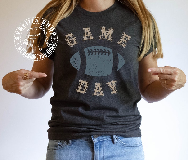 May include: A dark grey t-shirt with the text "GAME DAY" in a vintage style font. A distressed blue football graphic is centred on the shirt.