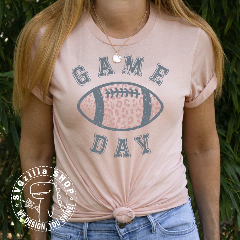 Pu&ograve; includere: Una maglietta rosa con una grafica grigia usurata di un pallone da calcio con un design a stampa leopardata rosa. Il testo "GAME DAY" &egrave; sopra il pallone.