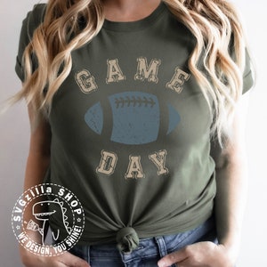 May include: A green t-shirt with a distressed vintage design featuring the text "GAME DAY" and a blue football graphic. The shirt is tied at the waist.