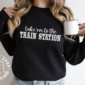 May include: Black sweatshirt with the text "take 'em to the TRAIN STATION" in white. A white dinosaur logo with the text "SVGzilla SHOP WE DESIGN, YOU SHINE!" is on the left sleeve.