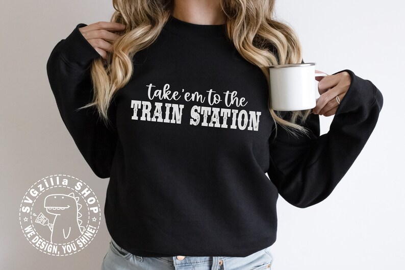 May include: Black sweatshirt with the text "take 'em to the TRAIN STATION" in white. A white dinosaur logo with the text "SVGzilla SHOP WE DESIGN, YOU SHINE!" is on the left sleeve.