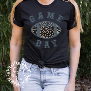 Pu&ograve; includere: Una maglietta nera con una grafica blu usurata di un pallone da calcio con un design a stampa leopardata. Il testo "GAME DAY" &egrave; scritto in un font blu usurato attorno al pallone da calcio.