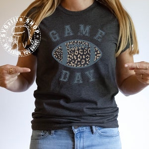 Pu&ograve; includere: Una maglietta grigio scuro con una grafica blu e bianca usurata di un pallone da calcio con un design a stampa leopardata. Il testo "GAME DAY" &egrave; stampato sotto il pallone da calcio.