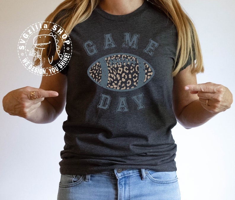 Pu&ograve; includere: Una maglietta grigio scuro con una grafica blu e bianca usurata di un pallone da calcio con un design a stampa leopardata. Il testo "GAME DAY" &egrave; stampato sotto il pallone da calcio.