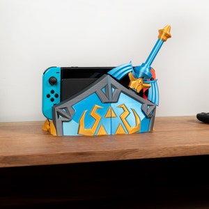 以下が含まれることがあります： ゼルダの伝説のビデオゲームシリーズの盾と剣の形をした青と金のビデオゲームコンソールスタンド。スタンドはプラスチック製で、Nintendo Switchコンソール用のスロットがあります。