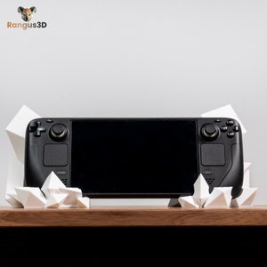 May include: A black handheld gaming console with a large screen and two controllers on the sides. The console is sitting on a white, geometric stand.