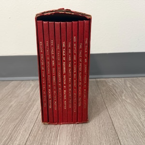 May include: A collection of red hardcover books, featuring titles like "The Tale of Peter Rabbit" and "The Tale of Squirrel Nutkin" by Beatrix Potter. The books are in a decorative red box, with the titles printed in white.