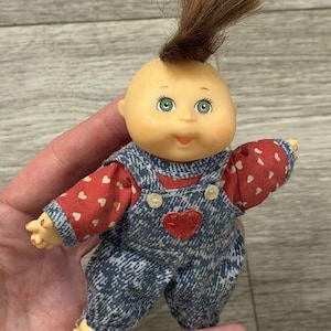 May include: A small doll with a round head, light skin, green eyes, and a single tuft of brown hair standing upright. The doll wears a red long-sleeved shirt with a white heart pattern under blue denim-style overalls featuring a red heart patch.