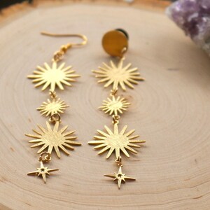 May include: Gold tone dangle earrings with a starburst design and a smaller star charm at the bottom. The earrings are on a wooden surface.