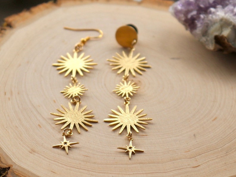 May include: Gold tone dangle earrings with a starburst design and a smaller star charm at the bottom. The earrings are on a wooden surface.